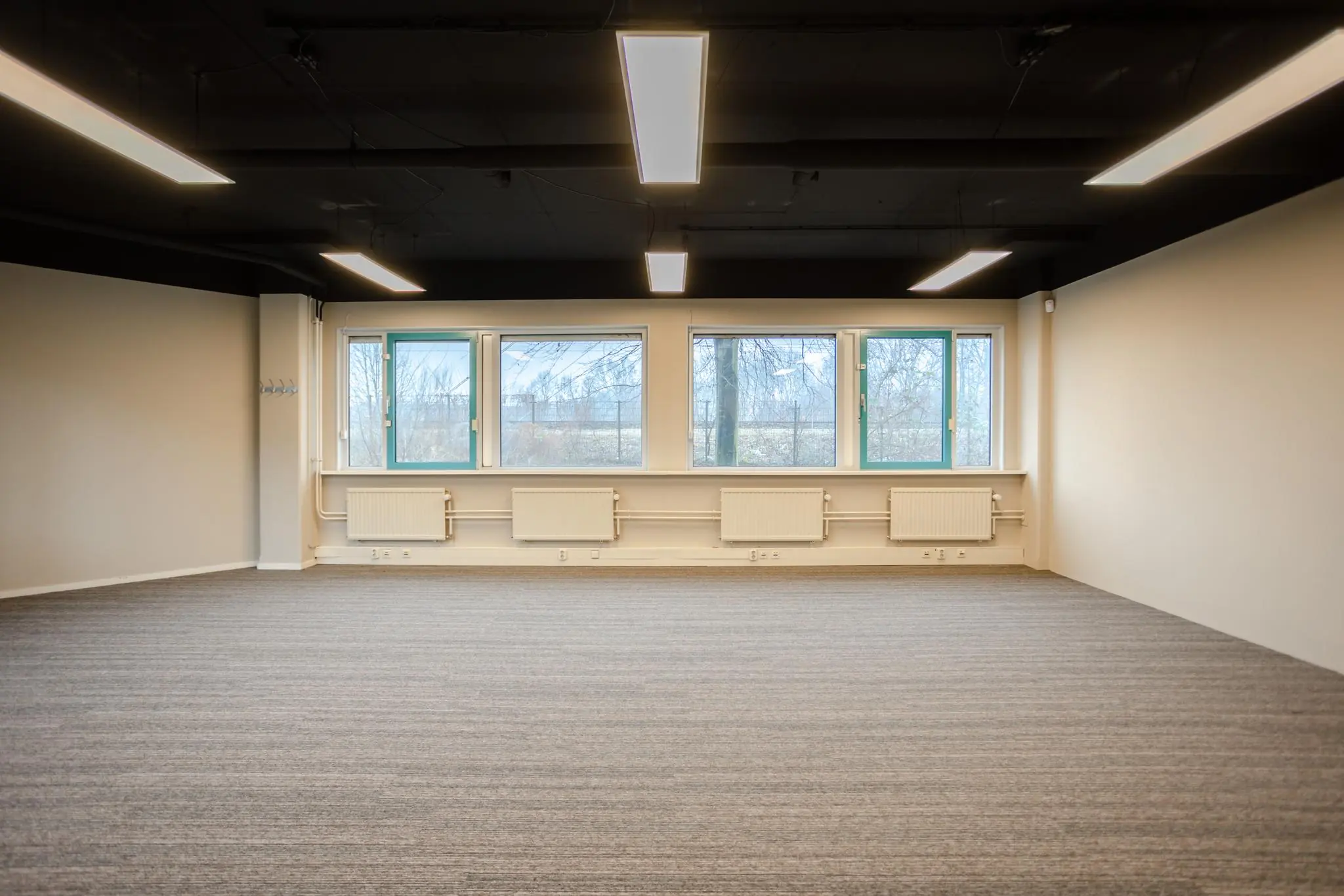 Leeg kantoor met brede ramen, grijze vloerbedekking en verlichting aan het zwarte plafond aan de Christiaan Krammlaan.